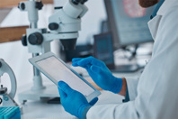 GettyImages-1487365113-scientist-with-gloves-tablet-in-hands-microscope-lab